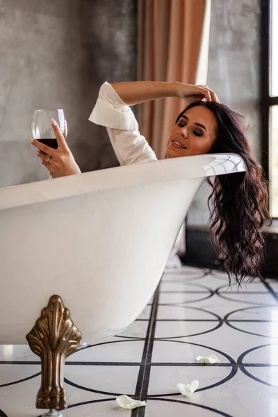 高級アパートでリラックスした赤ワインのゴブレットとレトロなバスルームに横たわっている幸せなファッション女性 — ストック写真