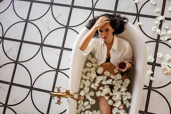 고전적 인 목욕 물 과하얀 장미꽃 잎 속에 누워 있는 매혹적 인 우아 한 갈색 머리 여인 — 스톡 사진