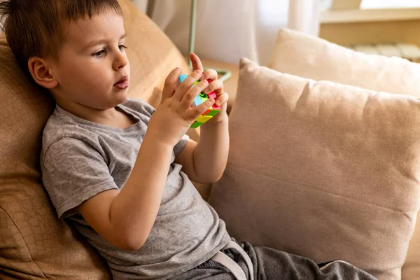 다양 한 색상의 루빅스 큐브를 조립하는 귀여운 사내 아이 집 소파에 누워 논리적 인 게임을 풀고 있습니다. — 스톡 사진