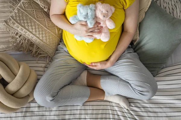 Вид зверху вагітна молода жінка тримає жовтий з двома милими ведмежими іграшками на животі, сидячи на ліжку Ліцензійні Стокові Зображення
