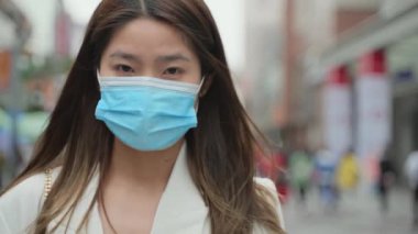 Asyalı genç iş kadınının yavaş çekimde çekilmiş portresi maske takıyor ve şehrin arka planındaki Coronavirus salgını konseptine bakıyor.