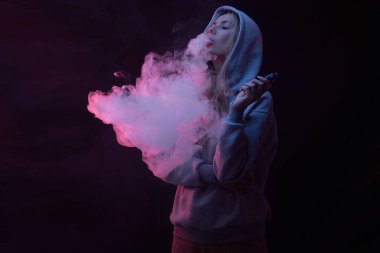 Gri kapüşonlu güzel sarışın kızın portresi siyah stüdyo arka planında izole edilmiş sigara içiyor, buhar dumanı bulutu, mini nargile.