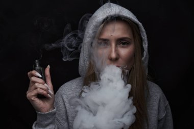 Gri kapüşonlu güzel sarışın kızın portresi siyah stüdyo arka planında izole edilmiş sigara içiyor, buhar dumanı bulutu, mini nargile.