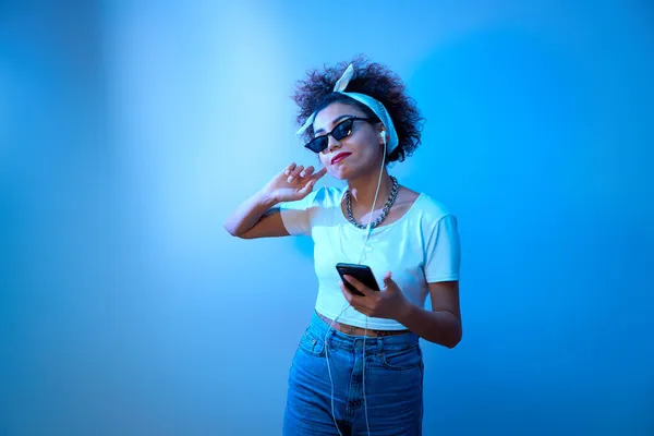 Trendy girl with afro curls listens and enjoys music with headphones in blue neon light, hip hop style model dances and relax
