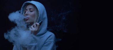 Gri kapüşonlu güzel sarışın kızın portresi siyah stüdyo arka planında izole edilmiş sigara içiyor, buhar dumanı bulutu, mini nargile.