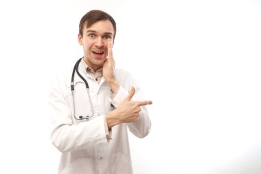Steteskoplu beyaz önlüklü yakışıklı doktor beyaz arka planda izole edilmiş boş bir alanı işaret ediyor. Ürünü gösteriyor, heyecan verici bir şey gösteriyor.