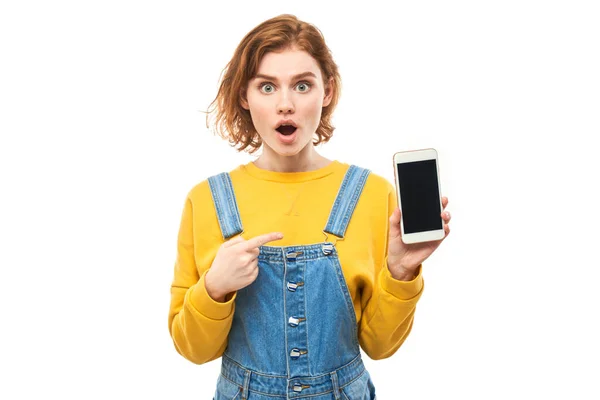喜怒无常的红头发女孩用手指指指智能手机空白屏幕 黄色衣服与白色工作室背景隔离 — 图库照片