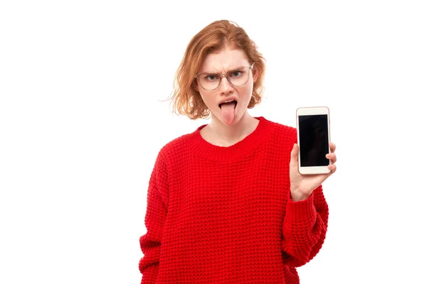 红头发女孩厌恶的脸展示了空白的智能手机屏幕 并展示了在白色工作室背景下隔离的眼镜和红色衣服的舌头 — 图库照片