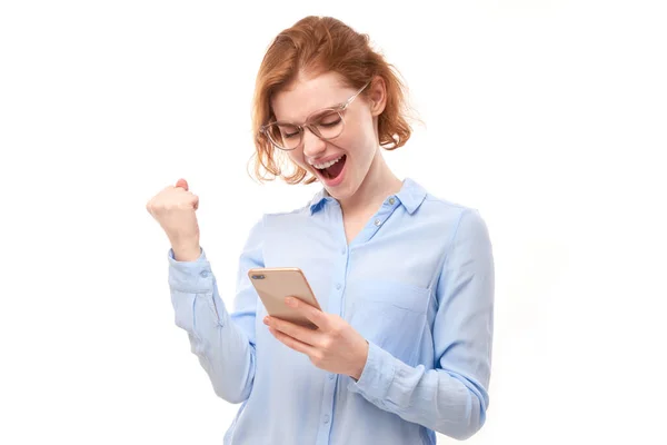 幸运的红头发女孩 身穿商务衬衫 戴着眼镜 隔离在工作室里 手持智能手机 收到了欢快的信息 庆祝胜利 — 图库照片