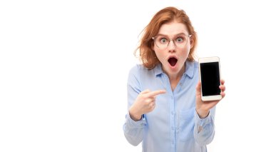 Şok olmuş kızıl saçlı kız beyaz stüdyo arka planında izole edilmiş gözlüklü ve iş gömlekli akıllı telefon boş ekranını işaret ediyor.