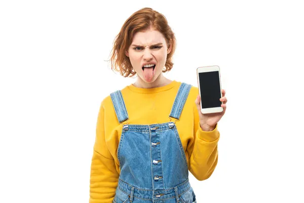 红头发女孩厌恶的脸展示了空白的智能手机屏幕 并展示了在白色工作室背景下隔离的黄色衣服的舌头 — 图库照片