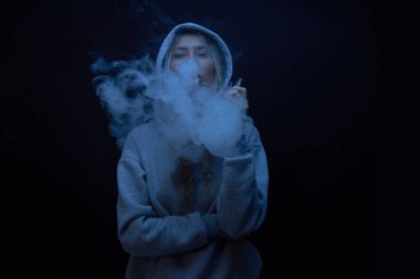 Gri kapüşonlu güzel sarışın kızın portresi siyah stüdyo arka planında izole edilmiş sigara içiyor, buhar dumanı bulutu, mini nargile.