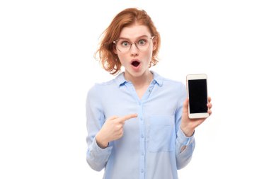 Şok olmuş kızıl saçlı kız beyaz stüdyo arka planında izole edilmiş gözlüklü ve iş gömlekli akıllı telefon boş ekranını işaret ediyor.
