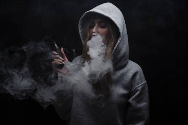 Gri kapüşonlu güzel sarışın kızın portresi siyah stüdyo arka planında izole edilmiş sigara içiyor, buhar dumanı bulutu, mini nargile.