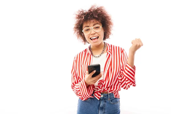 快乐地微笑着 带着智能手机的高加索女孩庆祝胜利 在白色的工作室背景上收到了快乐的信息 — 图库照片