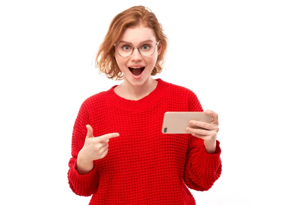 喜怒无常的红头发女孩把手指指向智能手机空白屏幕 眼镜和红色衣服被隔离在白色工作室的背景下 — 图库照片