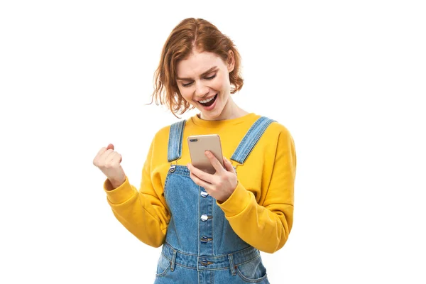 幸运的红头发女孩拿着智能手机 穿着黄色衣服 与白色工作室隔离 收到了欢快的信息 庆祝胜利 — 图库照片