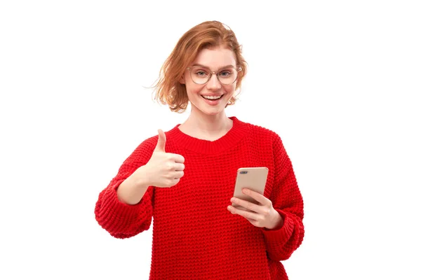 快乐的红头发女孩拿着智能手机 戴着眼镜 戴着红色衣服 与白色的工作室背景隔离在一起 — 图库照片