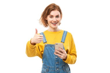 Happy redhead girl holding smartphone and thumbs up in yellow clothes isolated on white studio background