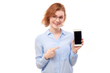 Şok olmuş kızıl saçlı kız beyaz stüdyo arka planında izole edilmiş gözlüklü ve iş gömlekli akıllı telefon boş ekranını işaret ediyor.