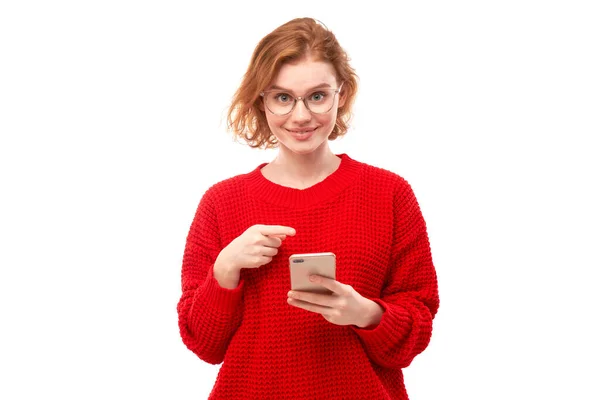 喜怒无常的红头发女孩把手指指向智能手机空白屏幕 眼镜和红色衣服被隔离在白色工作室的背景下 — 图库照片