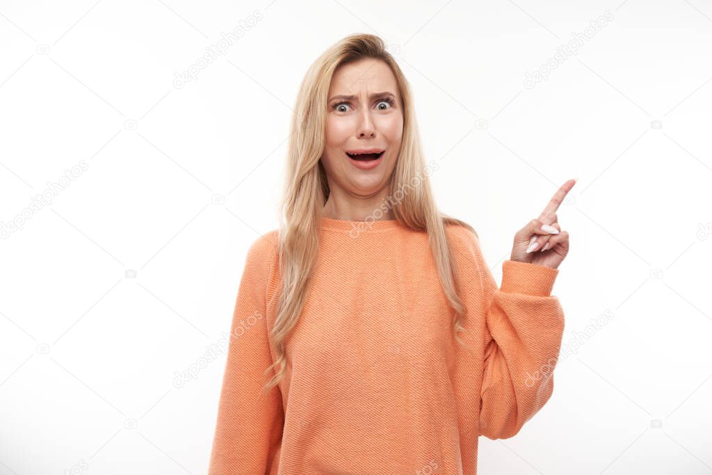 Shocked blond girl looks surprised on white studio background points finger at copy space