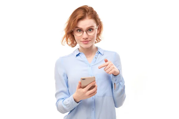 喜形于色的红头发女孩用手指指指智能手机的空白屏幕 眼镜和商务衬衫与白色工作室背景隔离在一起 — 图库照片