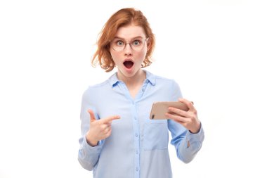 Şok olmuş kızıl saçlı kız beyaz stüdyo arka planında izole edilmiş gözlüklü ve iş gömlekli akıllı telefon boş ekranını işaret ediyor.