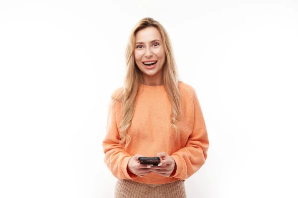穿着休闲装的漂亮金发姑娘望着智能手机屏幕 在白色的工作室背景下面带微笑 — 图库照片