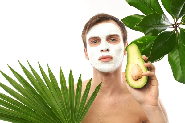 Portrait of young handsome man with cosmetic mask on his face holding avocado in hand isolated on exotic palm leaves background