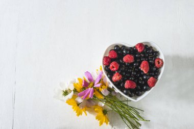 Beyaz kasede kırmızı ahududular ve kırmızı üzümler. Kalp şeklinde ve beyaz arka planda bir buket yabani yaz çiçeği.