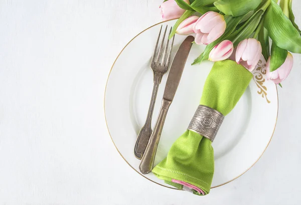 Bahar masa ayarı; pembe lale buketi, klasik çatal ve bıçak, beyaz tabak üst manzara üzerinde eski tip halka yeşil peçete