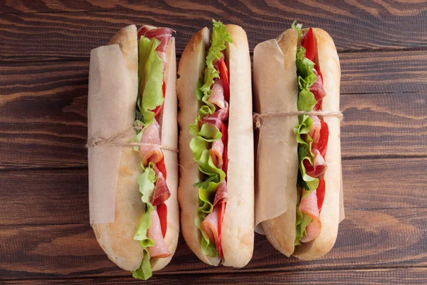 Üç banh mi sandviçi, dilimlenmiş domuz eti, biber ve ahşap arka planda kağıda sarılmış salata..