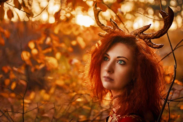 秋の森の鹿の角を持つ長い赤いドレスの女性. ロイヤリティフリーのストック画像
