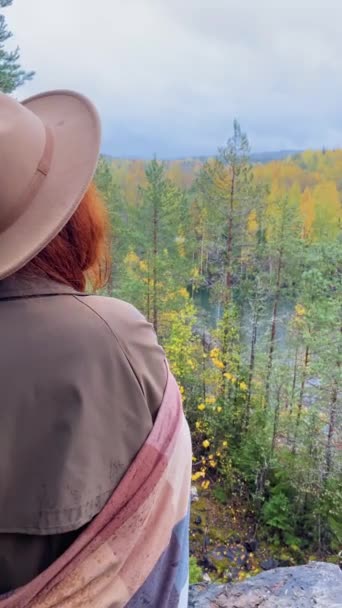 Red-haired girl in hat and raincoat stands on edge of cliff and looks at lake. — Stock Video