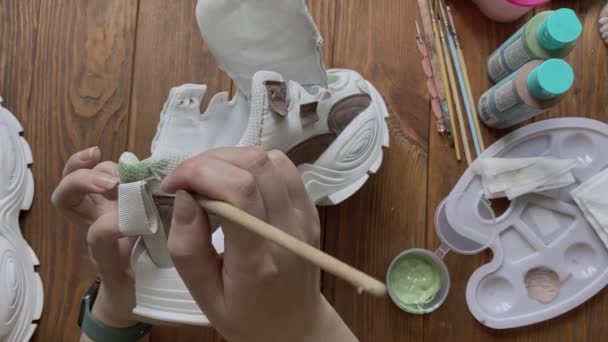 女孩们用手拿着画笔在木制桌子上画运动鞋的平铺 设计人员讲习班 在桌面上配备绿色丙烯酸和其他工具的调色板 业余爱好 创造力和家庭手工工作 — 图库视频影像