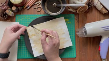 Dişiler el sanatları kağıdını su ve kahve solüsyonuyla süsler. Tasarımcı kâğıt koleksiyonu yapıp fırçayla boyar. Tabak, saç kurutma makinesi ve ahşap masadaki diğer aletler..