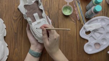 Top view of hands drawing on white boots with brush on desktop. Palette, acrylic paint in bottles and other tools on wooden table. Hobby, creativity and handmade work at home. Designers workshop.