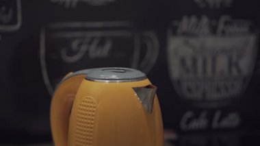 İnsanlar portakallı bir elektrikli su ısıtıcısının kapağını açar ve içine büyük bir şişeden temiz su döker. Ev aletleri mutfak masasında. Sıcak içecek yapmak için ev aletleri.