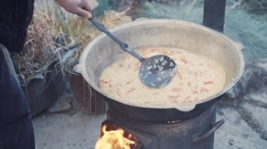 Person mixes pilaf in cauldron with slotted spoon. Cooking over campfire. Dish of Oriental cuisine made of rice, meat and vegetables. Charity lunch for poor. Plov in kazan smells delicious.