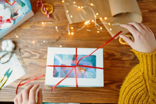 Flat lay of womans hands in orange knitted sweater opening gift box. New Years festive atmosphere. Christmas surprise with red ribbon and decorations on wooden table.