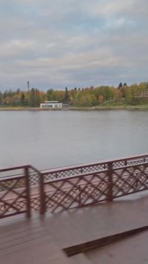 View from window of train departing from station. Rural platform with benches, lanterns and accompanying people. River, autumn forest and cloudy sky on background. Village landscape.