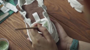 Artist applies green acrylic paint on white sneaker with brush. Tubes of paint and wooden table in background. Hobbies, handmade, crafts and leisure at quarantine.