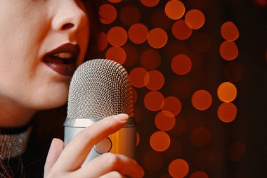 Close-up of womens lips painted with burgundy lipstick and retro microphone. Female singer with disco mic on bokeh light background. Redhead girl starting concert. Copy space for gig posters.