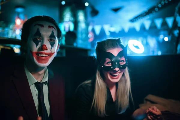 Woman in cat mask and guy with joker makeup sits at table in pub. Halloween party at a nightclub. All Saints Night. Garland lights on the background. Photo card with copy space.
