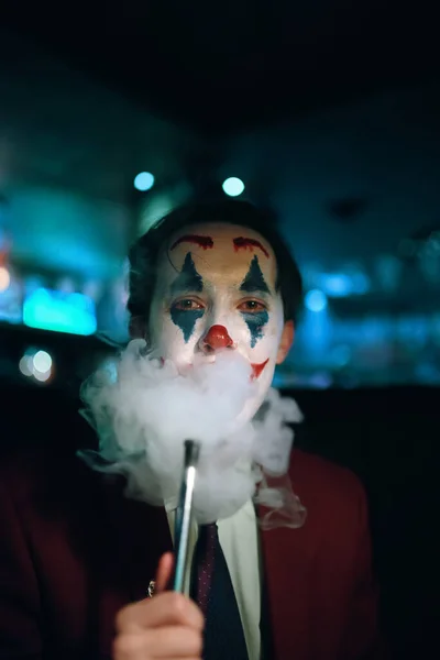 Guy in suit and tie with the Joker makeup puffs smoke out of his mouth. Autumn holiday. All Saints Night. Halloween party in nightclub with decorations and scary costumes.