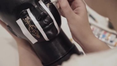 Kadın, rünleri mankenleri altın kalemle siyah kafaya boyuyor. Sanatçının çalışma masası. El işi ve hobi. Moda gösterisi ya da şapka vitrininin dekorasyonu için hazırlık.