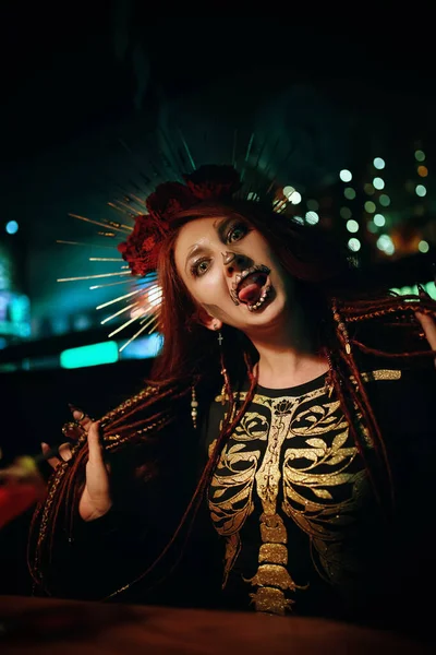 Female with skeleton makeup at a Halloween party make grimaces and looks at camera. Celebration in a nightclub. Woman in creepy costume of Mexican goddess of death. All Saints Night.