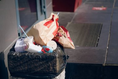 İçinde siyah çanta olan beton çöp kutusu. Bir deste kağıt paket, bir fincan kahve ve diğer içecekler çöpte. Fast food mağazasının yanındaki çöp tenekesi. Şehir manzarası.