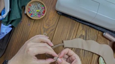 Kadın parmaklarının üst görüntüsü kayışı beyaz metal bir sürgüye bağlıyor. Ahşap bir masanın üzerinde işyeri. Hobi ya da el yapımı konsept. Terzi, eko-deriden yapılmış aksesuar veya etnik Slav başlığı dikiyor.